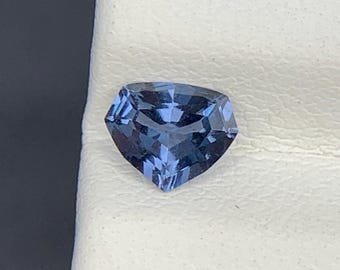 Pode incluir: Uma pedra preciosa solta com lapidação trillion, de tom azul-acinzentado profundo e superfície facetada. A pedra apresenta um formato triangular com cantos arredondados, exibindo reflexos de luz internos e uma textura polida e vítrea.