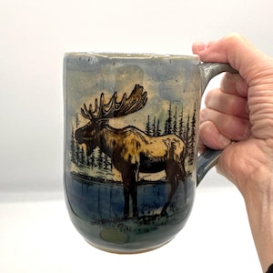 May include: A ceramic mug with a light blue and grey glaze featuring a detailed illustration of a brown moose standing near a dark blue lake with a forest of pine trees in the background. The mug has a sturdy handle and a smooth, rounded shape.