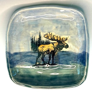 May include: Square glass dish with a painted landscape featuring a moose with large antlers, standing near a lake with a forest in the background. The dish has a light blue and green color scheme.