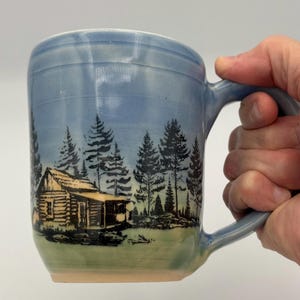 May include: A ceramic mug with a light blue glazed finish featuring a black ink illustration of a log cabin nestled among pine trees. The design wraps around the cylindrical body, which has a curved handle on the right side and a smooth rim.