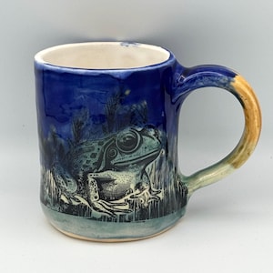 May include: A ceramic mug featuring a dark blue glaze with a black ink illustration of a frog sitting among tall grasses. The mug has a cream-colored interior and a curved handle that transitions from blue to a light tan color at the base.