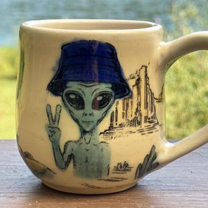 May include: A cream-colored ceramic mug featuring a blue-tinted alien wearing a dark blue bucket hat. The alien is making a peace sign gesture. The design includes a black line-art illustration of a desert landscape with rock formations.