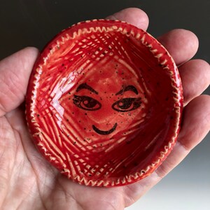 May include: A small, round, red ceramic dish featuring a hand-drawn black face with eyes, eyebrows, and a smile. The surface has a textured, cross-hatched pattern with small black speckles and a scalloped, ridged edge around the circular rim.