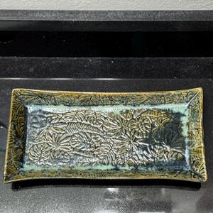 May include: A rectangular ceramic tray featuring a raised, embossed pattern of various tropical leaves across its surface. The glaze is a blend of deep forest green, olive, and dark blue tones, with a glossy finish that highlights the leaf textures.
