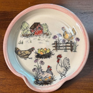 May include: A round ceramic spoon rest with a light cream base and a soft pink rim. The surface features a colorful illustration of a farm scene with a red barn, several chickens, a rooster, and nests with eggs surrounded by purple flowers.