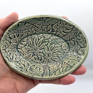 May include: An oval ceramic dish featuring a detailed, recessed botanical pattern of various tropical leaves. The surface is finished in a glossy, muted sage green glaze with dark, contrasting outlines highlighting the leaf textures.