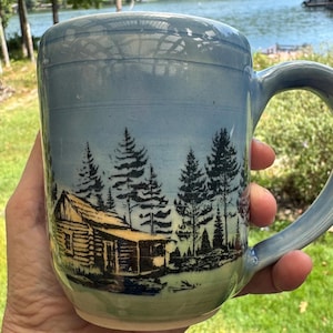 May include: A ceramic mug with a light blue and green gradient glaze featuring a black ink illustration of a log cabin nestled among tall pine trees. The mug has a smooth, rounded handle and a cylindrical body with visible horizontal wheel marks.