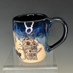 May include: A ceramic mug with a black handle and a dark blue interior. The exterior features a hand-drawn illustration of a haunted house, a black cat, and a tree. The mug has a cream-colored base and blue and white accents.