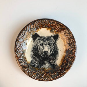 May include: A round ceramic dish featuring a detailed black and white illustration of a bear head in the center. The rim of the dish has a textured, dark brown glaze with an intricate, carved pattern that contrasts with the smooth, cream-colored center.