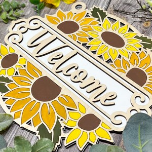 May include: Wooden welcome sign with a white background and the word "Welcome" in a decorative font. The sign is decorated with multiple yellow sunflowers with brown centres and green leaves.