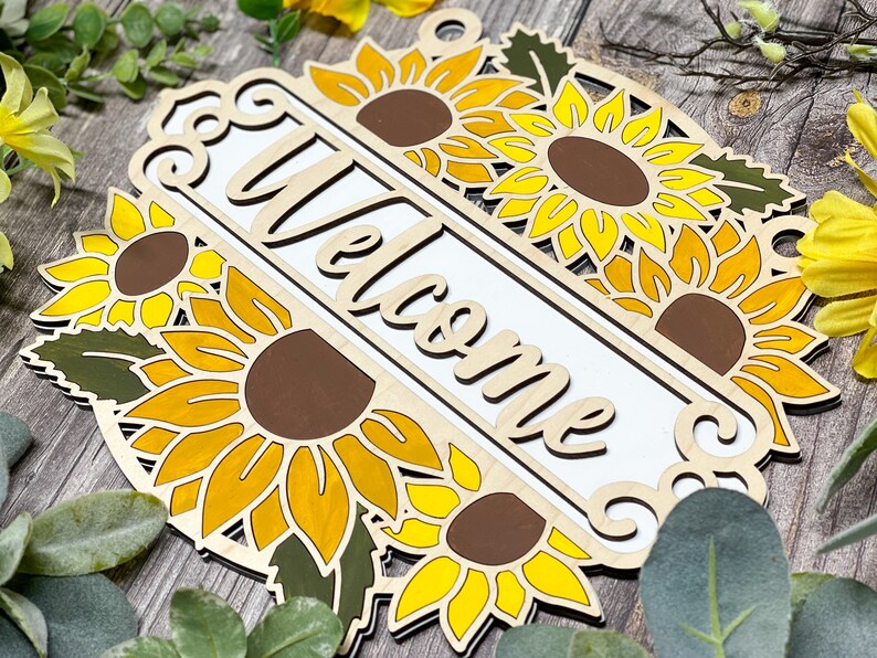 May include: Wooden welcome sign with a white background and the word "Welcome" in a decorative font. The sign is decorated with multiple yellow sunflowers with brown centres and green leaves.