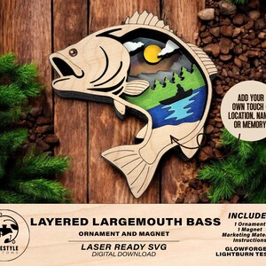 May include: Layered wooden ornament of a largemouth bass with a scenic lake scene inside. The ornament is laser cut and ready to be assembled. The text "Layered Largemouth Bass Ornament and Magnet" is at the top of the image. The text "Laser Ready SVG Digital Download" is below the bass. The text "Includes 1 Ornament 1 Magnet Marketing Materials Instructions Glowforge & Lightburn Tested!" is on the right side of the image.