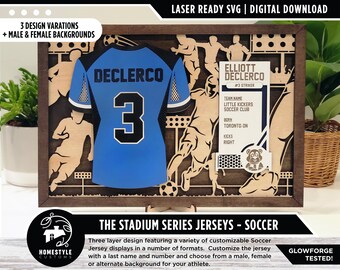 May include: A laser-cut wooden frame with a blue soccer jersey inside. The jersey has the name "Declerco" and the number "3" on the front. The frame also includes a player's information card with the name "Elliott Declerco", the team name "Little Kickers Soccer Club", and the player's number "3". The frame is part of a series called "The Stadium Series Jerseys - Soccer".