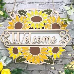 May include: Wooden welcome sign featuring a sunflower design. The sign is painted in shades of yellow and brown and displays the word "Welcome" in a decorative font.