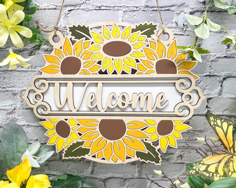 May include: Wooden welcome sign featuring a sunflower design. The sign is painted in shades of yellow and brown and displays the word "Welcome" in a decorative font.