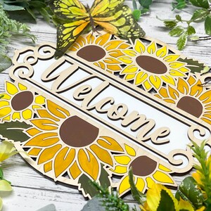 May include: Wooden welcome sign featuring a sunflower design. The sign is painted in shades of yellow, brown, and green. The word "Welcome" is written in white on the sign.