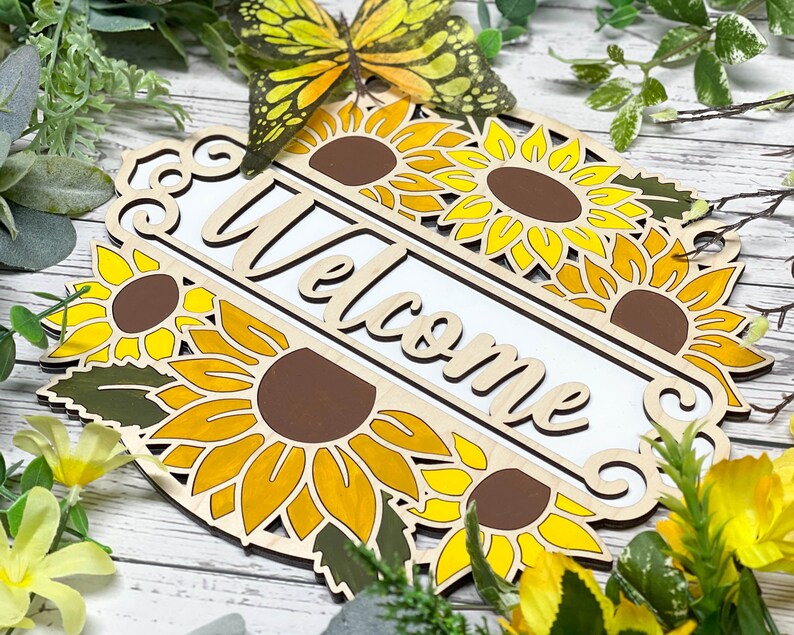 May include: Wooden welcome sign featuring a sunflower design. The sign is painted in shades of yellow, brown, and green. The word "Welcome" is written in white on the sign.