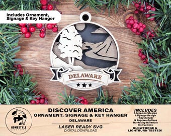 Pode incluir: Ornamento, sinalização e porta-chaves de madeira cortados a laser, apresentando uma imagem estilizada de uma árvore e um rio com o texto "DELAWARE" e três estrelas. O design faz parte de uma série "Discover America".