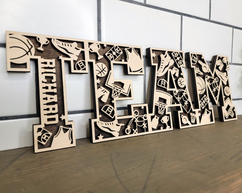 Peut inclure: Lettres en bois &eacute;pell&eacute;es "TEAM" sur le th&egrave;me du basketball. Chaque lettre est d&eacute;cor&eacute;e de d&eacute;coupes de ballons de basket, de maillots, de troph&eacute;es et d'autres images li&eacute;es au basketball. La lettre "T" comprend &eacute;galement le nom "Richard" en orientation verticale.