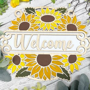 May include: Wooden welcome sign featuring a sunflower design. The sign is painted white with the word "Welcome" in a decorative font. The sunflowers are yellow with brown centres.
