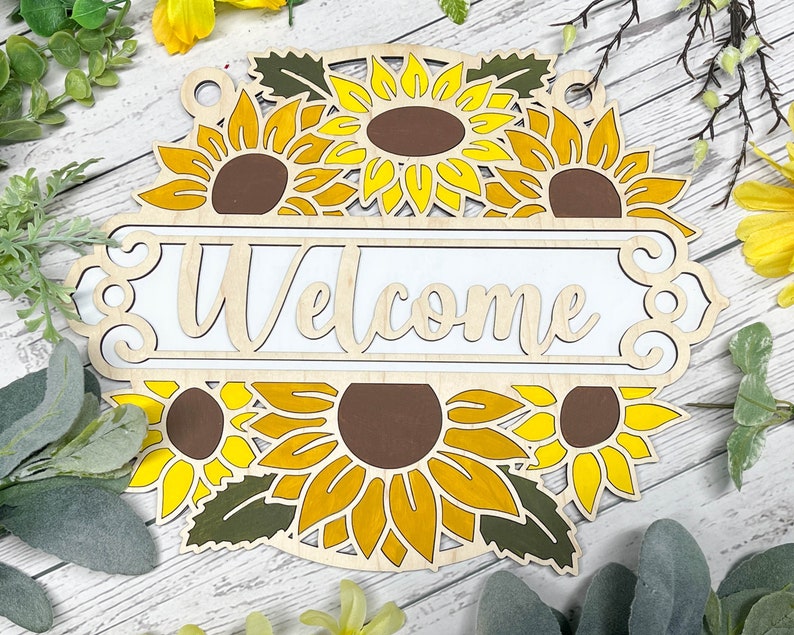 May include: Wooden welcome sign featuring a sunflower design. The sign is painted white with the word "Welcome" in a decorative font. The sunflowers are yellow with brown centres.
