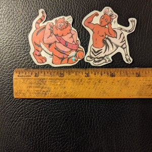 May include: Two colorful stickers featuring a tiger and a zebra with human-like bodies. The tiger is orange and black with a red mane and a blue sash. The zebra is white and black with a pink mane and a blue tail.