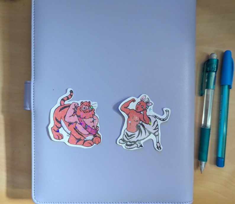 May include: Two colorful stickers featuring anthropomorphic animal characters. The first sticker is a tiger with orange and black stripes, wearing a red and white sash. The second sticker is a zebra with black and white stripes, wearing a red sash and a crown.