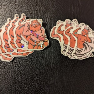 May include: A set of five stickers featuring a tiger and a zebra. The tiger is orange and black with a red mane and is flexing its muscles. The zebra is white and black with a red mane and is flexing its muscles. The stickers are all on a black background.