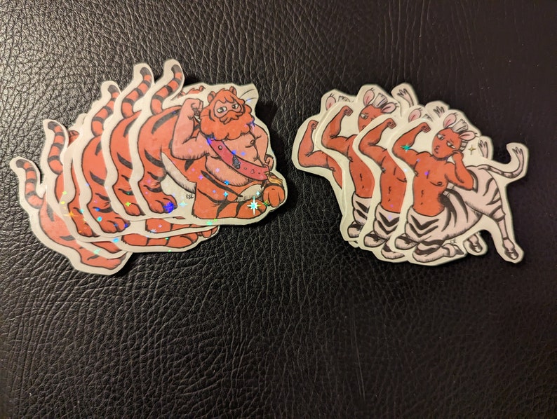 May include: A set of five stickers featuring a tiger and a zebra. The tiger is orange and black with a red mane and is flexing its muscles. The zebra is white and black with a red mane and is flexing its muscles. The stickers are all on a black background.
