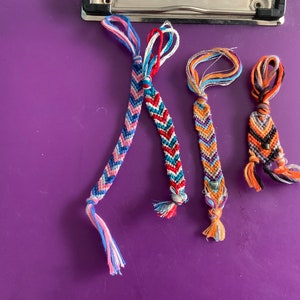 Może przedstawiać: Trzy kolorowe bransoletki przyjaźni z wzorami w chevron. Bransoletki są wykonane z nici w różnych kolorach i mają frędzle na końcach.