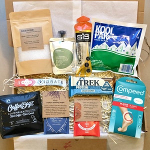 May include: A brown cardboard box filled with various items, including a bag of bath salts labelled "Restore", a bottle of energy drink labelled "Go Energy", a protein bar labelled "Trek", a bag of tea labelled "Night Time", a bag of coffee labelled "Coffee Bagz", a package of instant ice packs labelled "Kool Pak", a package of blister plasters labelled "Compeed", and a card with the text "Especially for you".