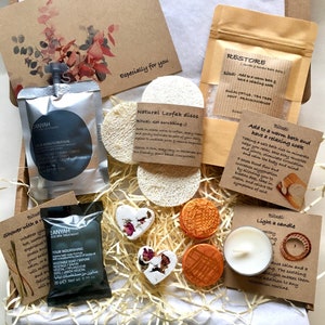 May include: A gift box filled with bath and body products, including a natural loofah sponge, bath salts, a heart-shaped bath bomb, a candle, and a small bottle of body cream. The box is lined with brown paper and straw. The products are labelled with the words "Restore", "Natural Loofah Discs", "Add to a warm bath and have a relaxing soak", "Shower with a Ritual", "Light a candle", and "Your Nourishing Vegetable Soap".