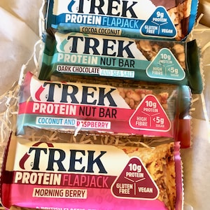 May include: Four Trek protein bars in a box. The top bar is blue Cocoa Coconut flapjack, followed by a green Dark Chocolate and Sea Salt nut bar, a pink Coconut and Raspberry nut bar, and a bottom pink Morning Berry protein flapjack.