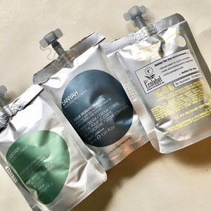 May include: Three silver pouches with blue and green labels. The labels contain text in English and Arabic. The pouches are labeled "Anyah Eco Spa Treatment" and contain body care products. The pouches are 1.01 fluid ounces each.