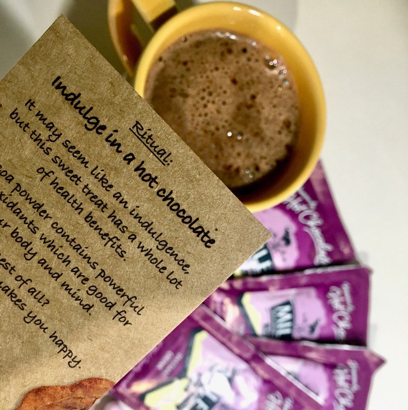 May include: A brown paper card with the text "Ritual: Indulge in a hot chocolate" and "It may seem like an indulgence, but this sweet treat has a whole lot of health benefits. Cocoa powder contains powerful antioxidants which are good for your body and mind. Best of all? Makes you happy."  The card is partially covering a yellow mug filled with hot chocolate.  There are several purple packets of hot chocolate mix in the background.