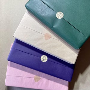 May include: Four wrapped gifts stacked on top of each other. The top gift is wrapped in green paper with a "Merry Christmas" sticker. The second gift is wrapped in beige paper with a heart-shaped sticker. The third gift is wrapped in purple paper with a "Happy Birthday" sticker. The bottom gift is wrapped in pink paper with a heart-shaped sticker.