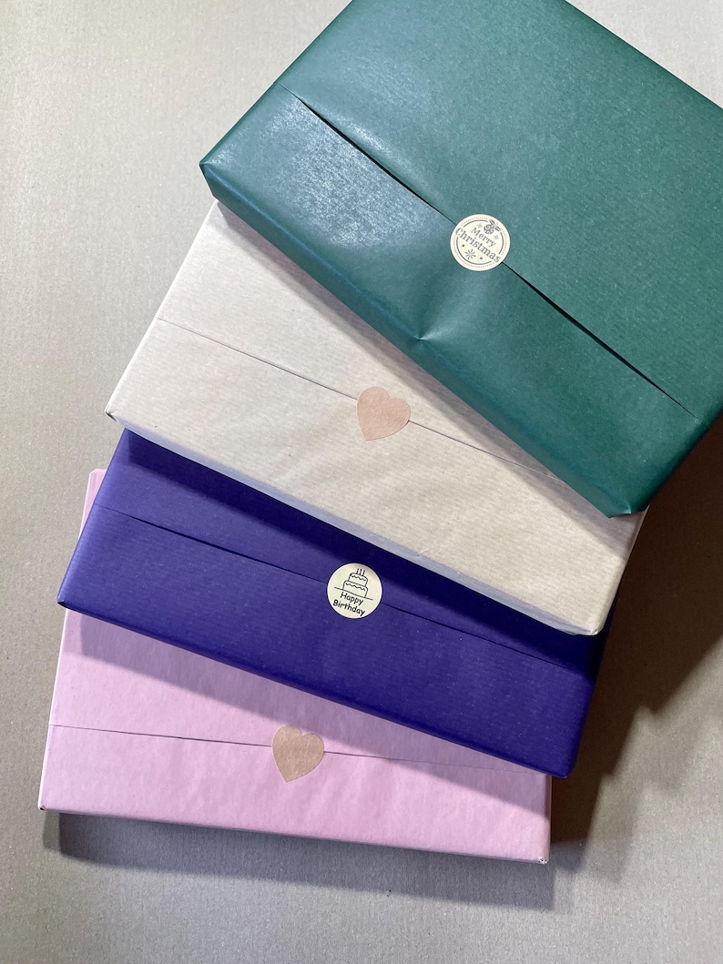 May include: Four wrapped gifts stacked on top of each other. The top gift is wrapped in green paper with a "Merry Christmas" sticker. The second gift is wrapped in beige paper with a heart-shaped sticker. The third gift is wrapped in purple paper with a "Happy Birthday" sticker. The bottom gift is wrapped in pink paper with a heart-shaped sticker.