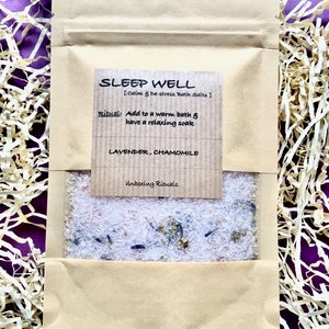 May include: A brown paper pouch with a label that reads "SLEEP WELL [Calm & De-stress Bath Salts]". The label also includes the text "Ritual: Add to a warm bath & have a relaxing soak" and "LAVENDER, CHAMOMILE". The pouch contains white bath salts with lavender and chamomile flowers.