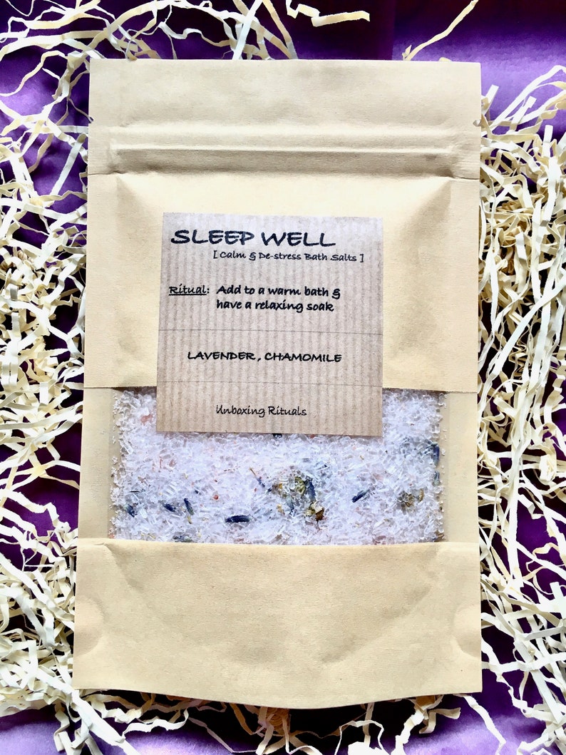 May include: A brown paper pouch with a label that reads "SLEEP WELL [Calm & De-stress Bath Salts]". The label also includes the text "Ritual: Add to a warm bath & have a relaxing soak" and "LAVENDER, CHAMOMILE". The pouch contains white bath salts with lavender and chamomile flowers.