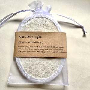 May include: An oval-shaped natural loofah sponge with white fabric trim, enclosed in a sheer white drawstring pouch. A brown paper label is wrapped around the centre, printed with the text: Natural Loofah, Ritual: Get scrubbing :), Eco-friendly body care.