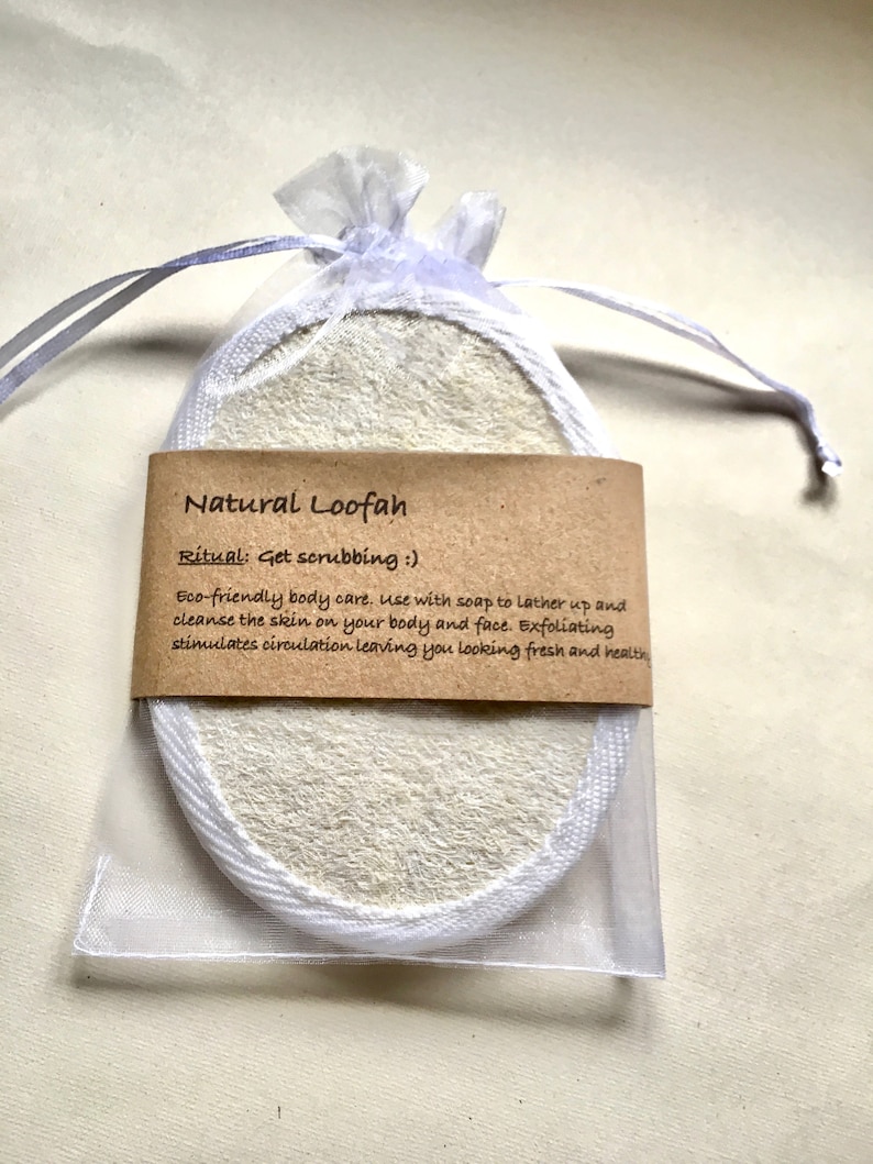 May include: An oval-shaped natural loofah sponge with white fabric trim, enclosed in a sheer white drawstring pouch. A brown paper label is wrapped around the centre, printed with the text: Natural Loofah, Ritual: Get scrubbing :), Eco-friendly body care.