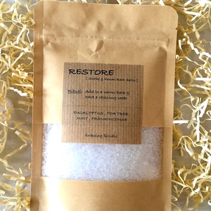 May include: A brown paper bag filled with white bath salts. The bag has a label that says "RESTORE [Soothe & Renew Bath Salts]" and lists the ingredients: Eucalyptus, Tea Tree, Mint, Frankincense. The label also says "Ritual: Add to a warm bath & have a relaxing soak" and "Unboxing Rituals".