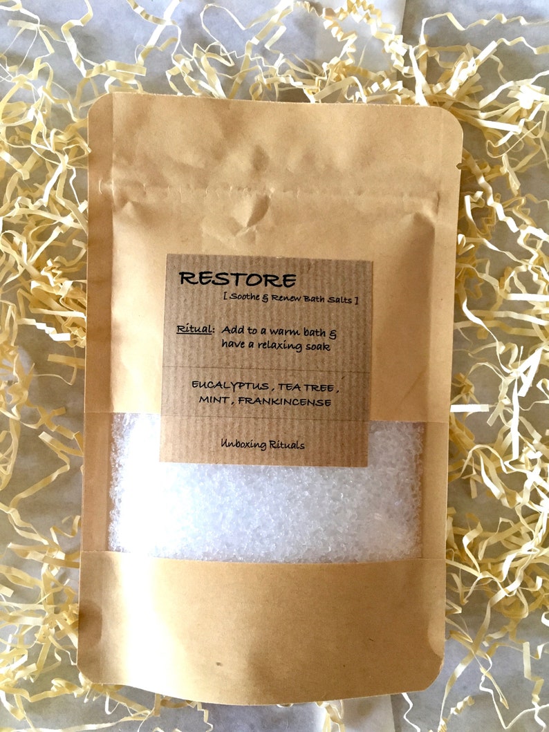 May include: A brown paper bag filled with white bath salts. The bag has a label that says "RESTORE [Soothe & Renew Bath Salts]" and lists the ingredients: Eucalyptus, Tea Tree, Mint, Frankincense. The label also says "Ritual: Add to a warm bath & have a relaxing soak" and "Unboxing Rituals".