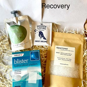 May include: A self-care gift set containing blister plasters, shower gel, bath salts, and tea. The box is filled with straw. The packaging includes the words "Recovery", "Sweet Dreams", and "Restore".