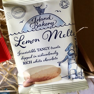 May include: A white and blue package of Island Bakery Lemon Melts. The package features a cartoon illustration of a cow on a hill with the text "Island Bakery Lemon Melts". The text "Irresistible TANGY treats, dipped in ridiculously RICH white chocolate" is also on the package.