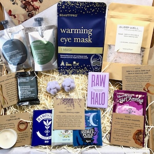 May include: A gift box filled with self-care items, including a warming eye mask, bath salts, tea bags, hot chocolate, and a candle. The box is filled with shredded paper and has a purple and blue color scheme.