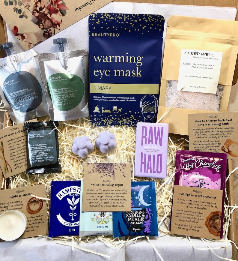 May include: A gift box filled with self-care items, including a warming eye mask, bath salts, tea bags, hot chocolate, and a candle. The box is filled with shredded paper and has a purple and blue color scheme.