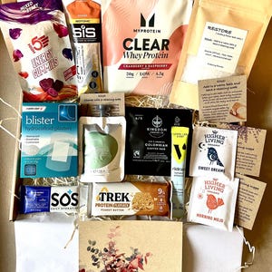 May include: A selection of wellness products presented in a gift box. Contents include protein powder, energy gummies, blister plasters, coffee, tea bags, a protein flapjack, and hydration tablets. A card with floral design is also included.
