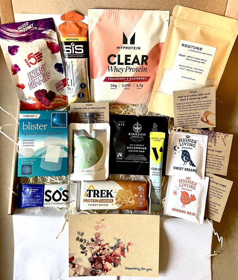 May include: A selection of wellness products presented in a gift box. Contents include protein powder, energy gummies, blister plasters, coffee, tea bags, a protein flapjack, and hydration tablets. A card with floral design is also included.