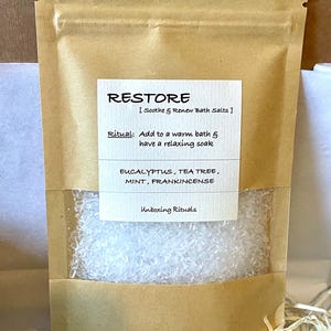 May include: A brown paper bag containing white bath salts, with a clear window. The label reads "RESTORE" and lists ingredients and instructions. The text "Unboxing Rituals" is also visible on the label.